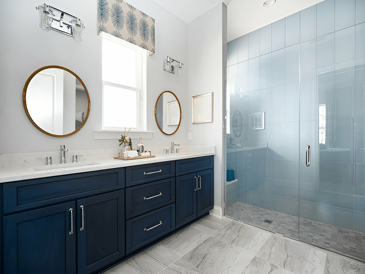 Modern bathroom with navy blue double vanity, round mirrors, white countertops, and large glass shower.