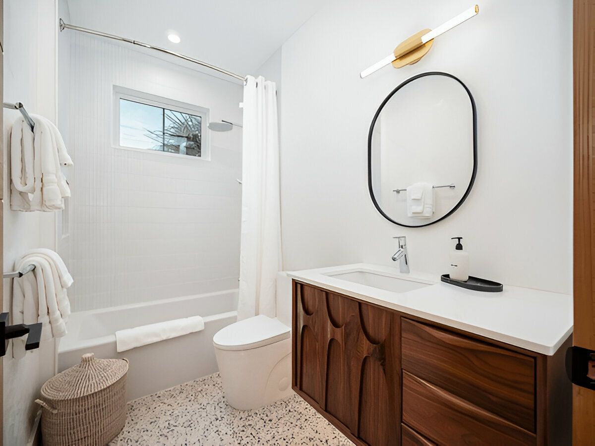 Modern bathroom with wood vanity, terrazzo floor, oval mirror, and white bathtub with shower curtain.