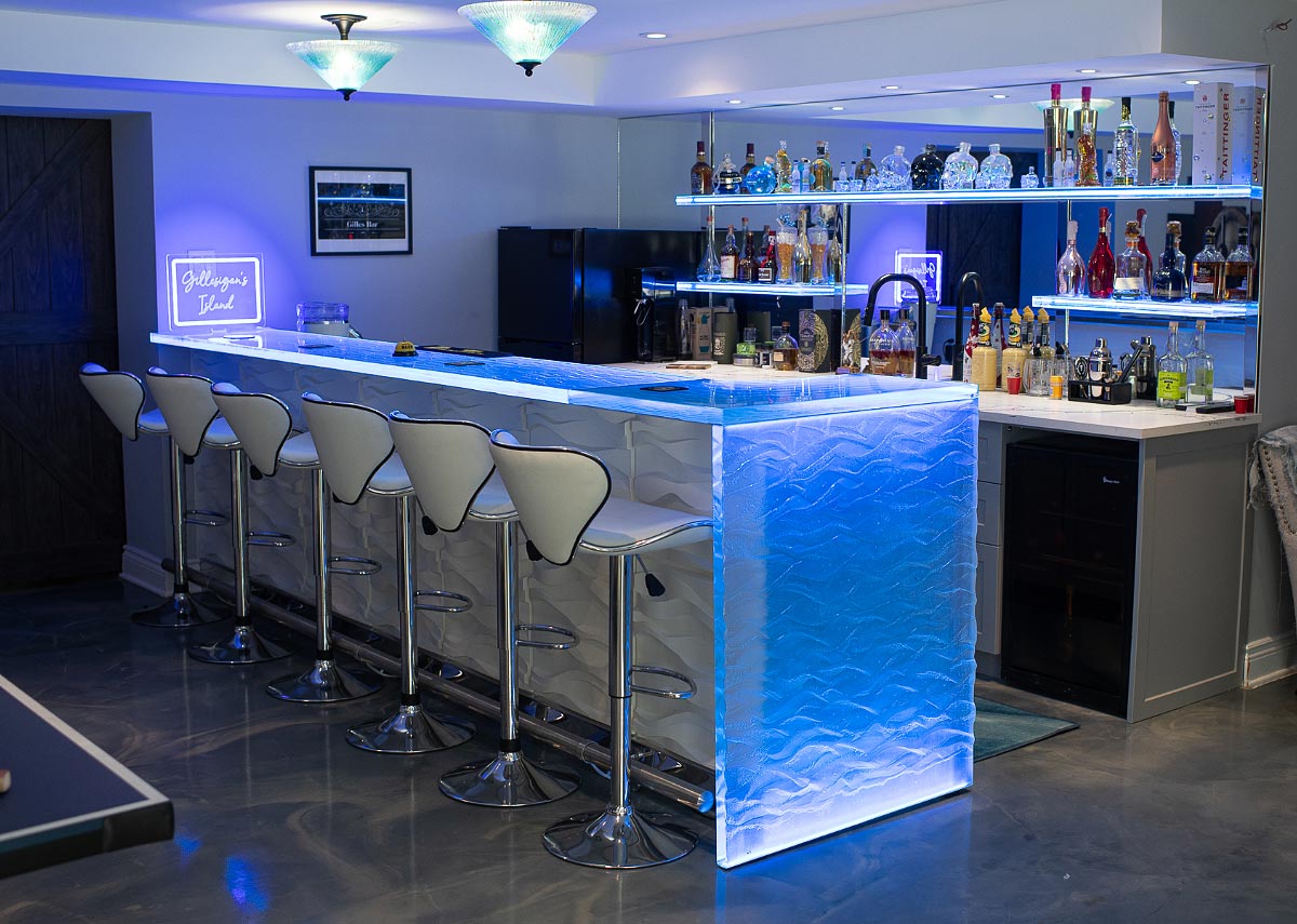 Contemporary home bar with illuminated LED counter, white bar stools, glass shelving, and liquor display