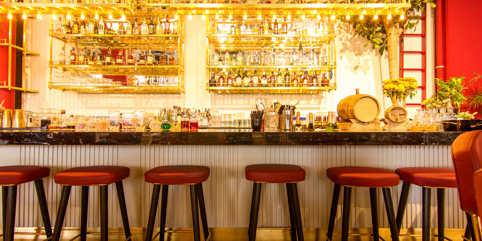 Elegant bar setup with golden liquor shelves, black marble countertop, red cushioned bar stools, and ambient lighting