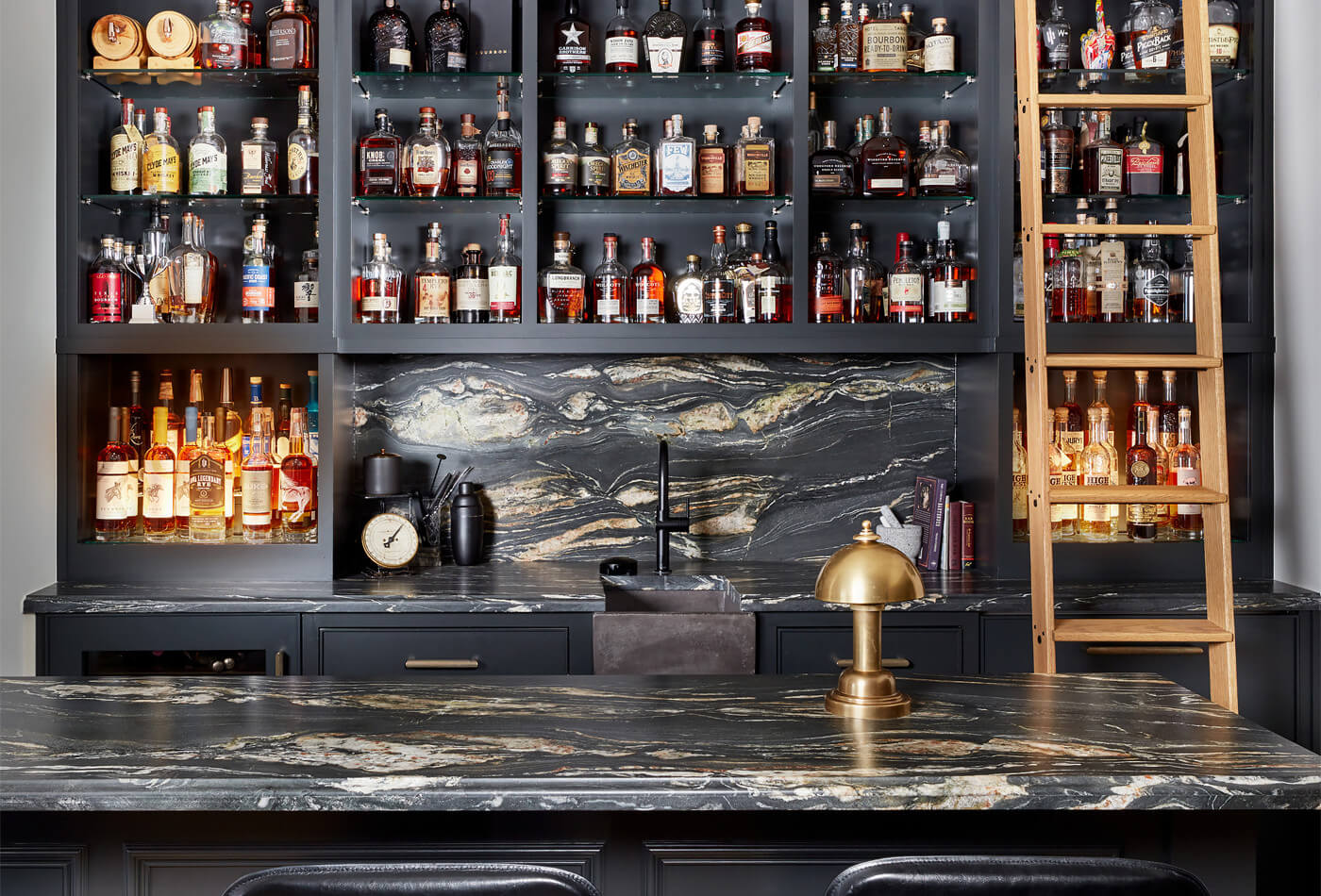 Luxurious home bar featuring a dark veined marble countertop and backsplash, matte black cabinetry, built-in liquor display shelves, a gold desk lamp, and a wooden library ladder