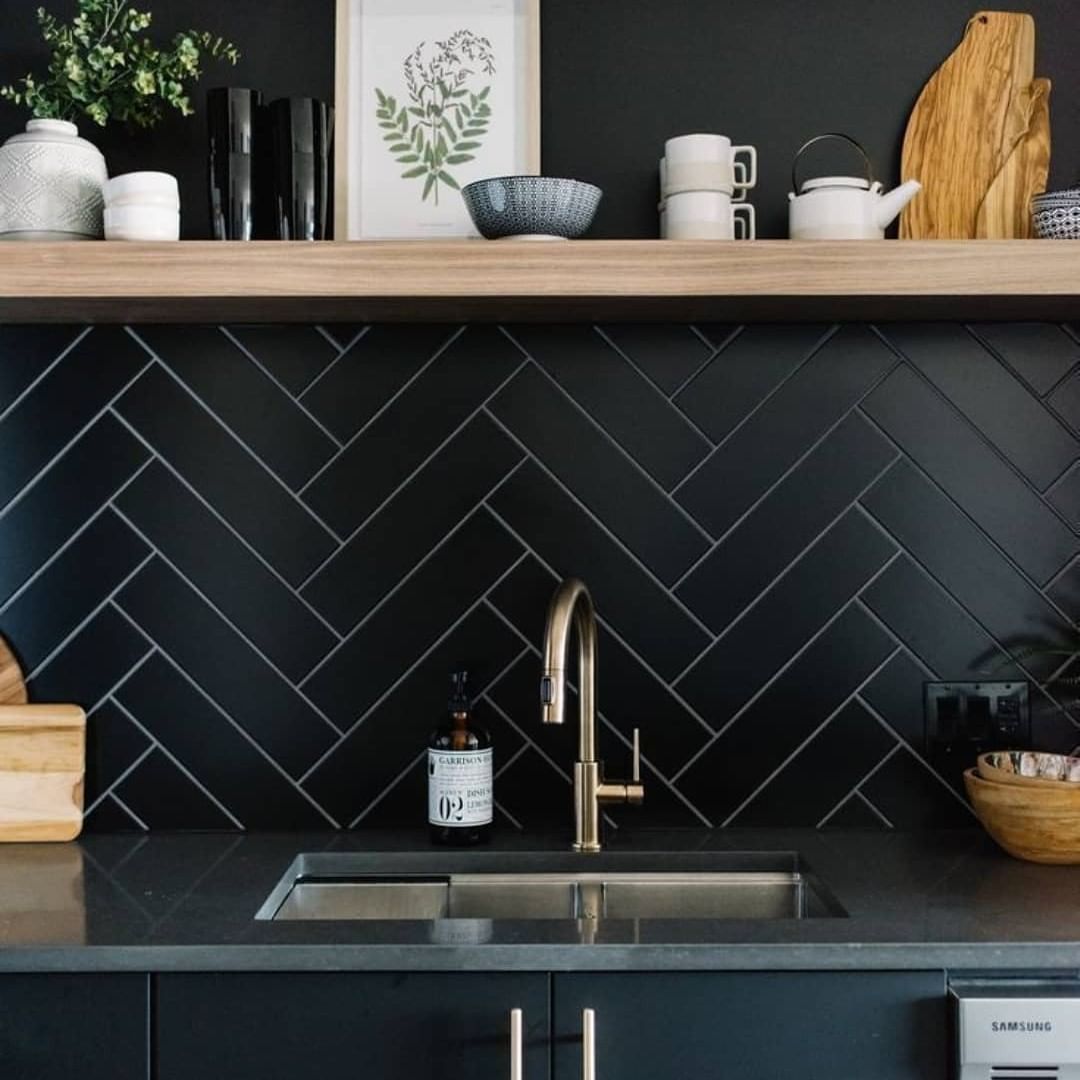 Contemporary kitchen featuring matte black herringbone tile backsplash, brass faucet, gray countertop, and wooden open shelf