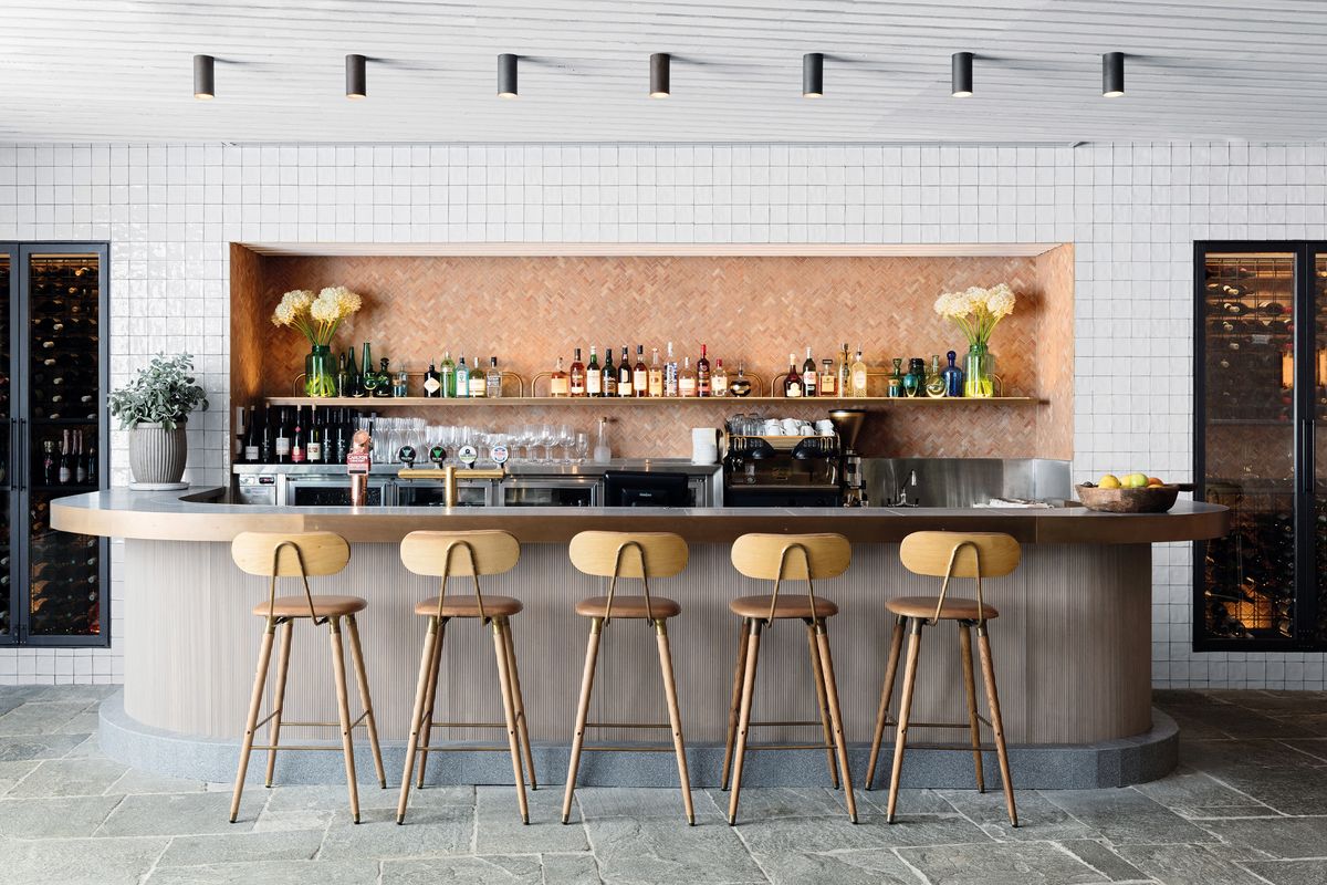 Contemporary bar with light wood counter, Scandinavian-style bar stools, copper backsplash, open shelves with liquor bottles and glassware, and minimalist lighting