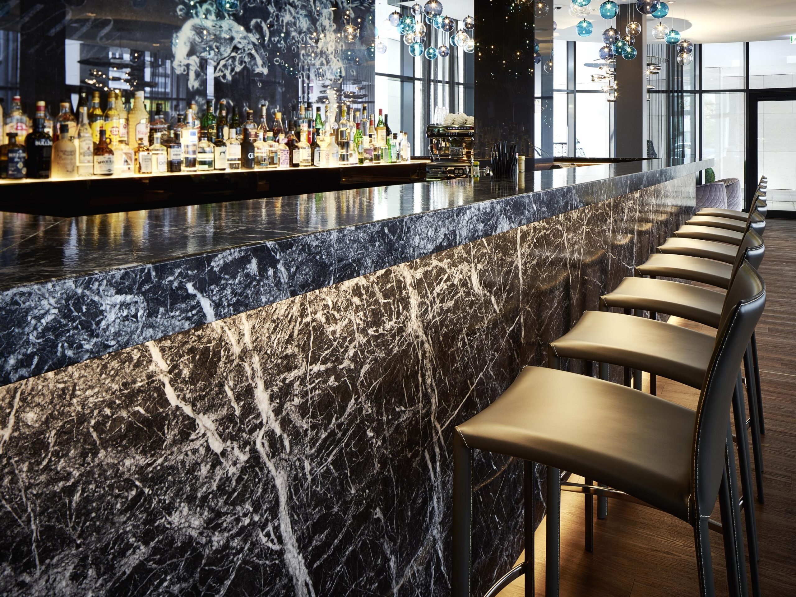 Elegant bar with black marble countertop and front, modern high-back leather bar stools, illuminated liquor display, and decorative hanging lights in a high-end lounge