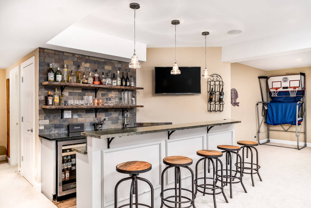 Home bar setup featuring a stone backsplash, floating wood shelves stocked with glassware and bottles, a marble countertop, pendant lighting, and a nearby indoor basketball game