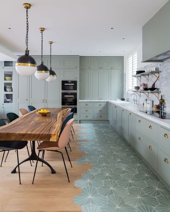 Modern kitchen with sage green cabinetry, a live-edge wood dining table, and geometric patterned tiles transitioning into light wood flooring.