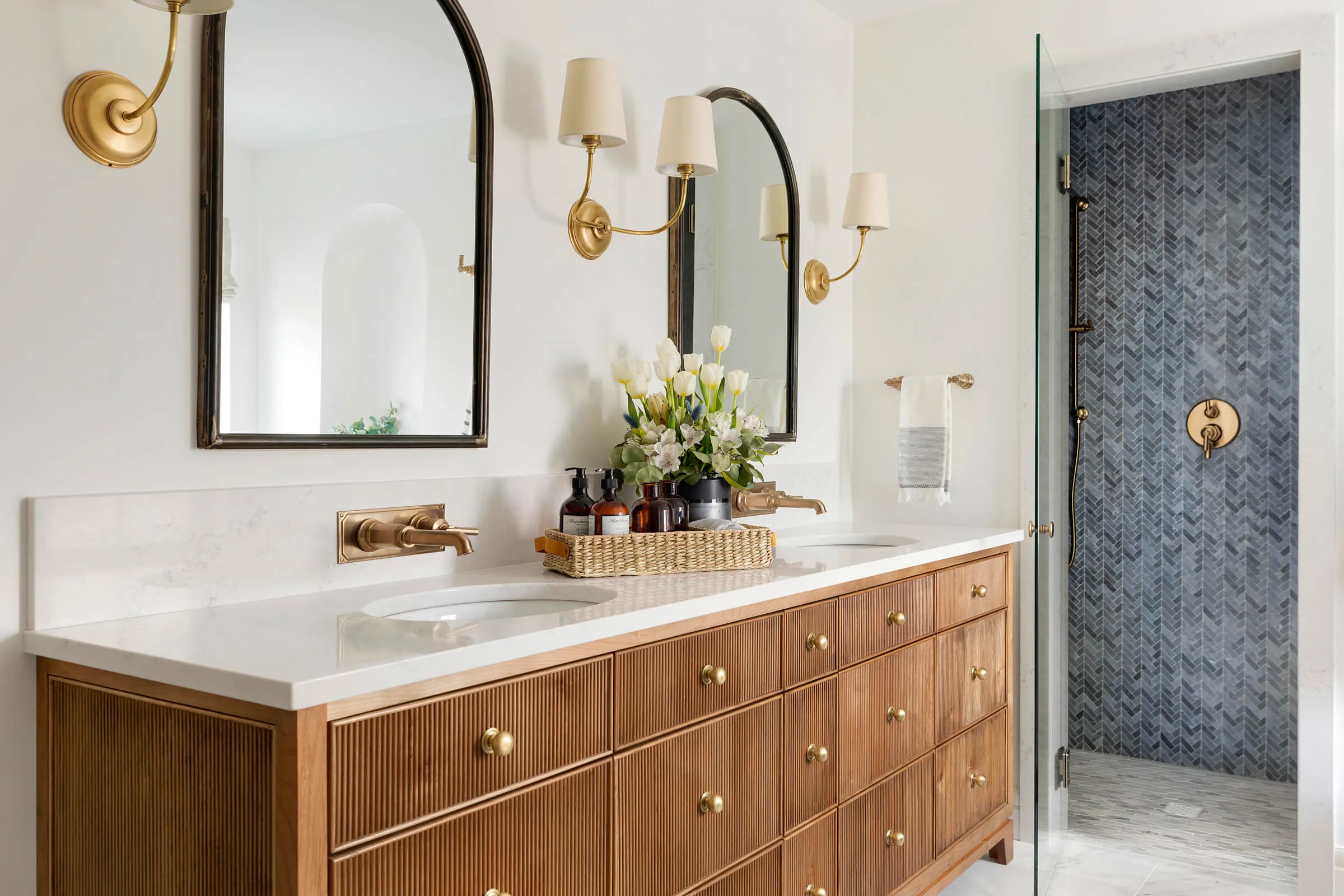 Double-sink bathroom vanity with fluted wood drawers and gold accents, next to navy chevron tile shower