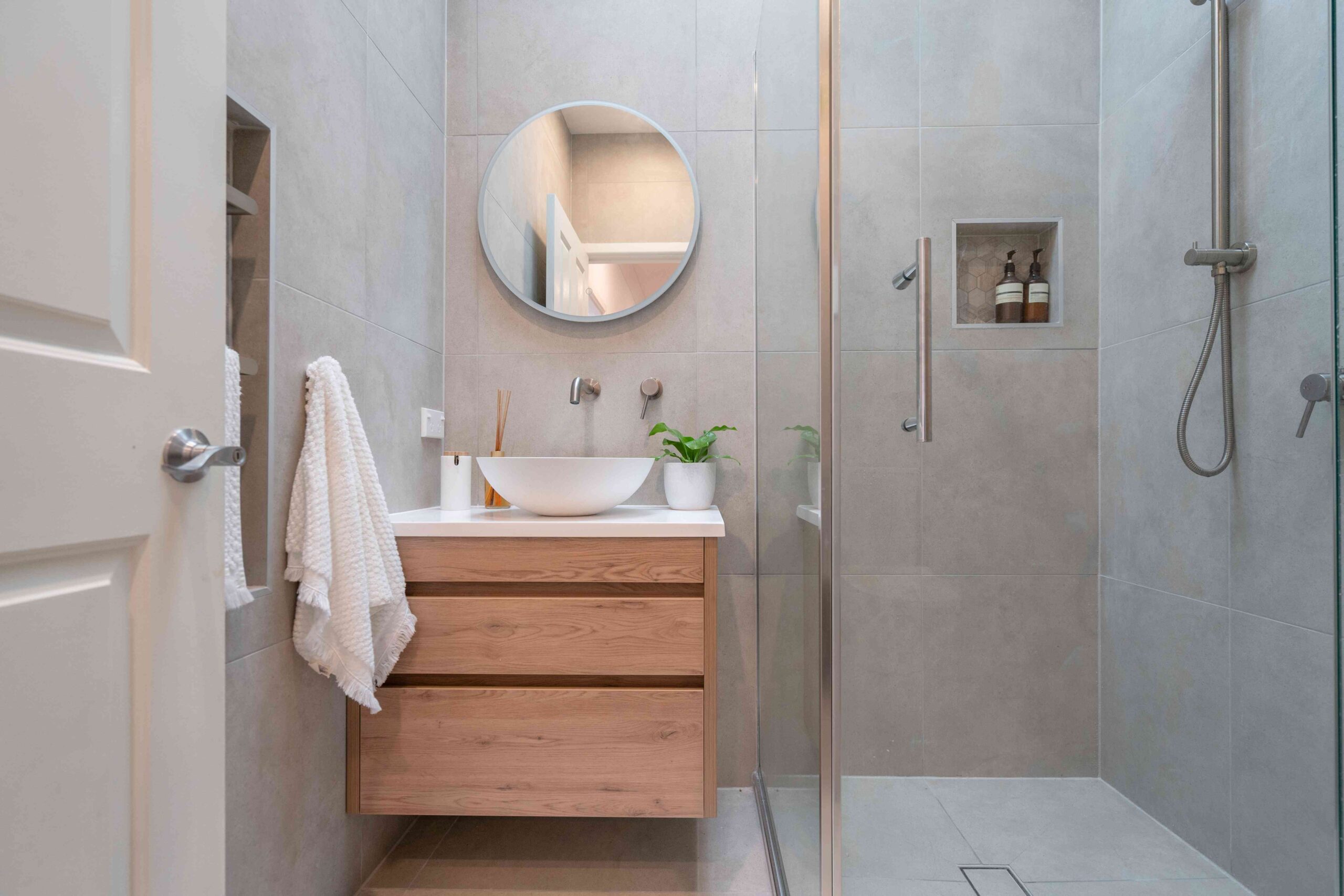 Minimalist bathroom with light wood vanity, round mirror, wall-mounted faucet, and a walk-in shower with built-in niche