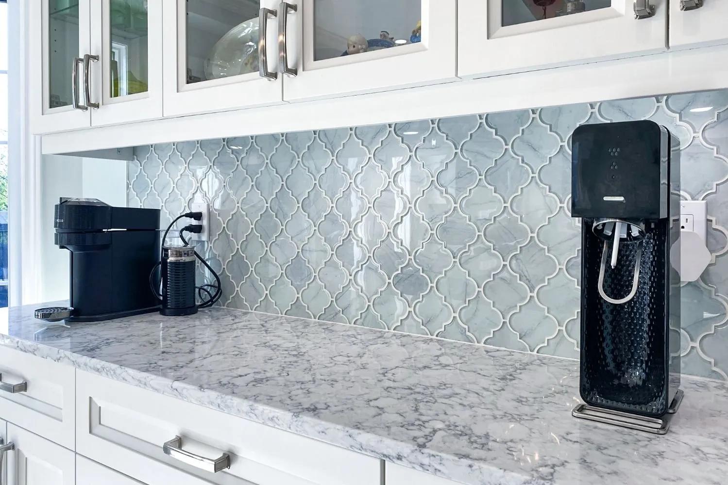 Sleek kitchen with light blue arabesque tile backsplash and white marble countertop under glass-front cabinets.