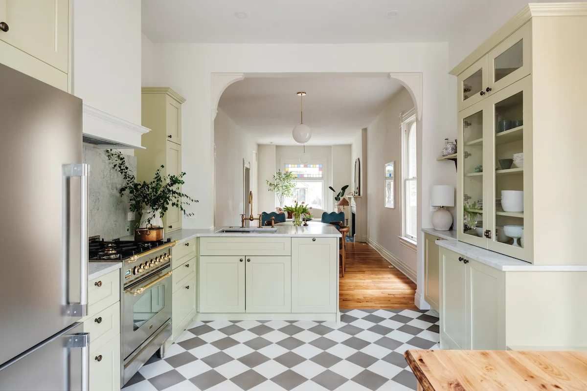 Kitchen with soft sage green cabinetry, brass hardware, marble countertops, and a gray-and-white checkerboard tile floor, leading to a light-filled living room.