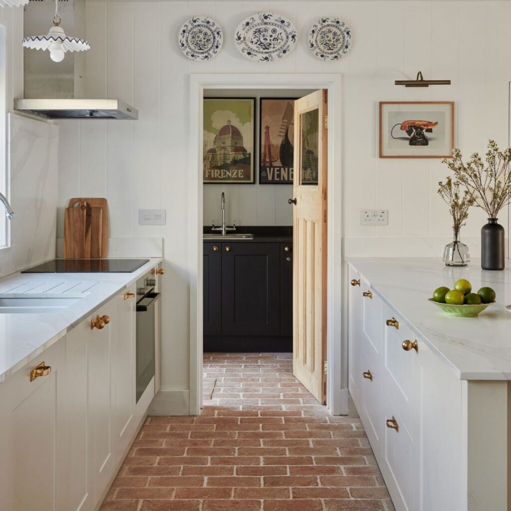 A cozy white kitchen with gold hardware, marble countertops, and red brick tile flooring leading into a pantry with black cabinetry and vintage travel posters.