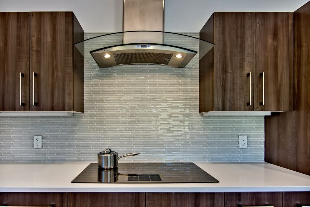Contemporary kitchen with sleek glass tile backsplash, walnut wood cabinets, a black electric cooktop, and a stainless steel range hood.