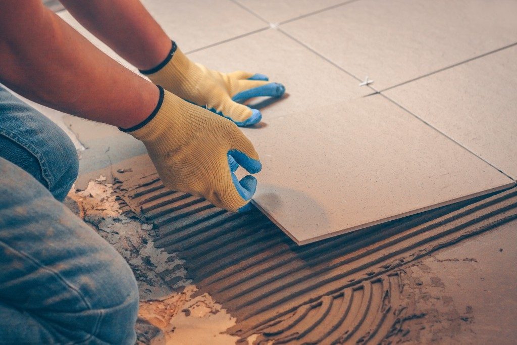 Close-up of a construction worker installing ceramic floor tiles using adhesive mortar and wearing protective gloves.