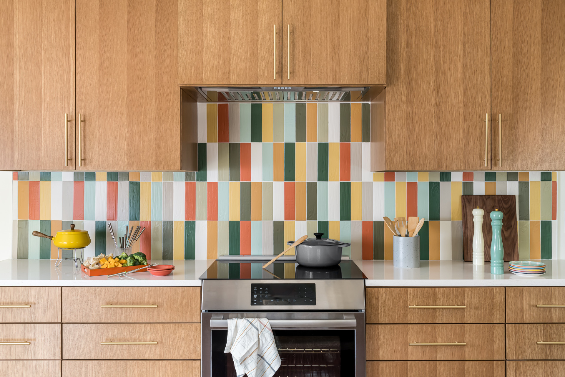 Bright and cheerful kitchen featuring a vertical multicolor tile backsplash, natural wood cabinets, and a white countertop with cooking accessories.