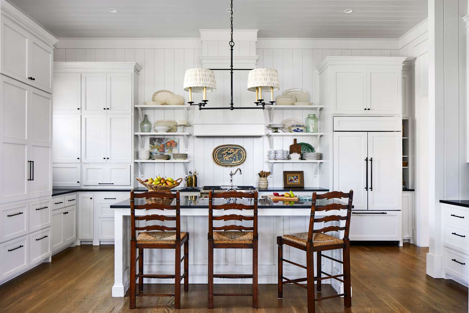 Traditional white kitchen with open shelving, wooden barstools, and black countertops.