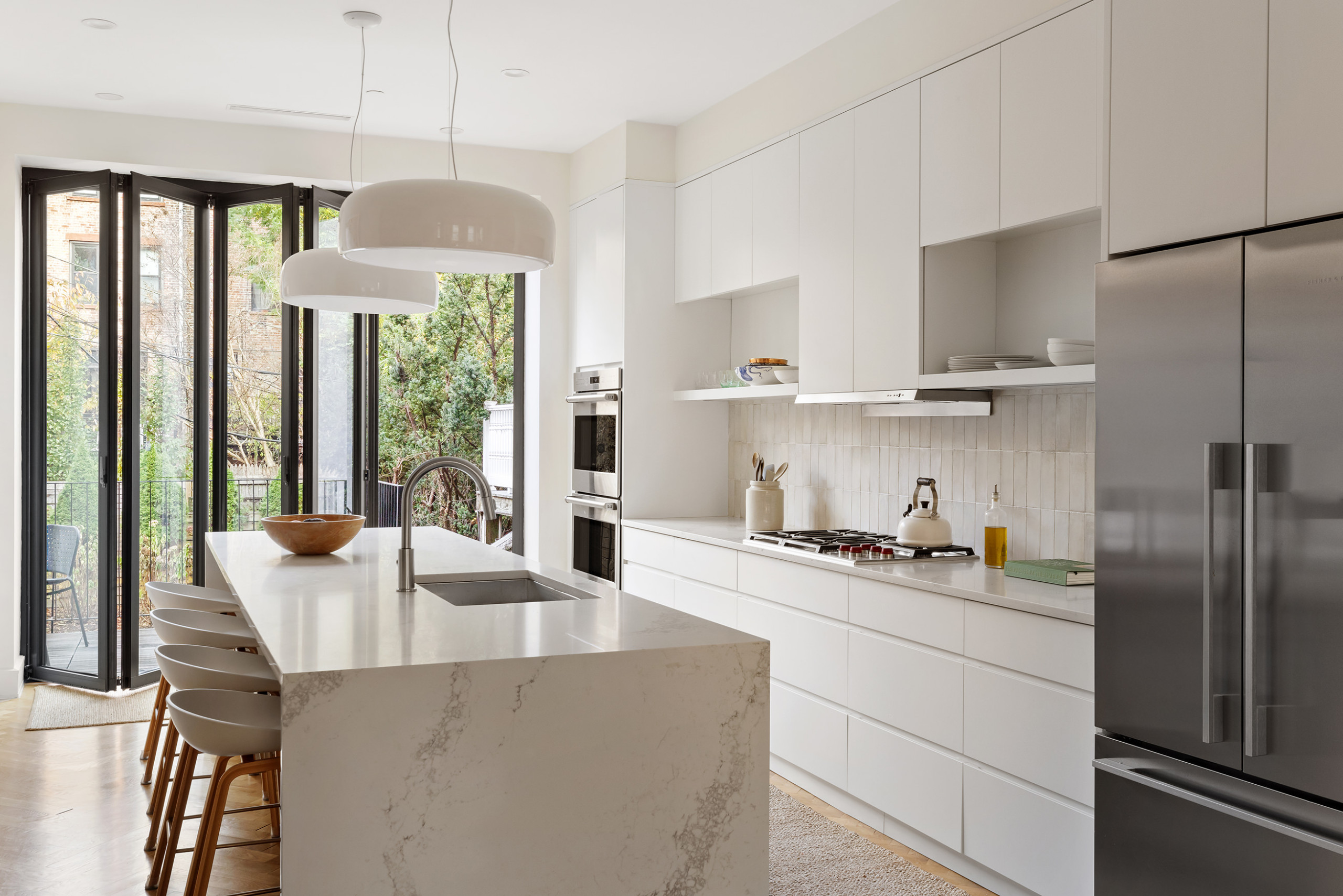 Modern white kitchen with marble island, sleek cabinetry, and large glass doors opening to a green backyard.