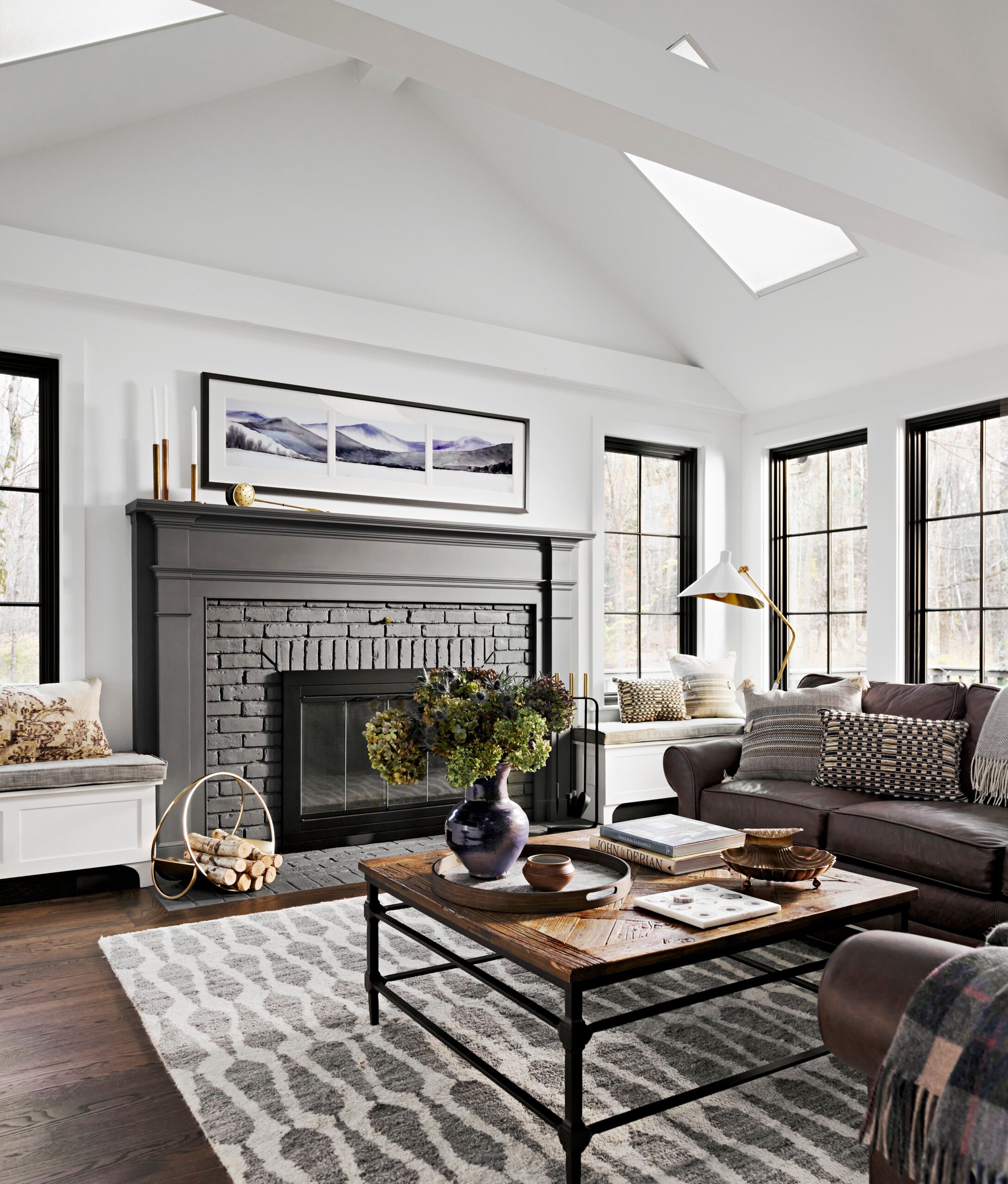 Contemporary living room with black brick fireplace, brown leather sofa, rustic coffee table, and large windows.