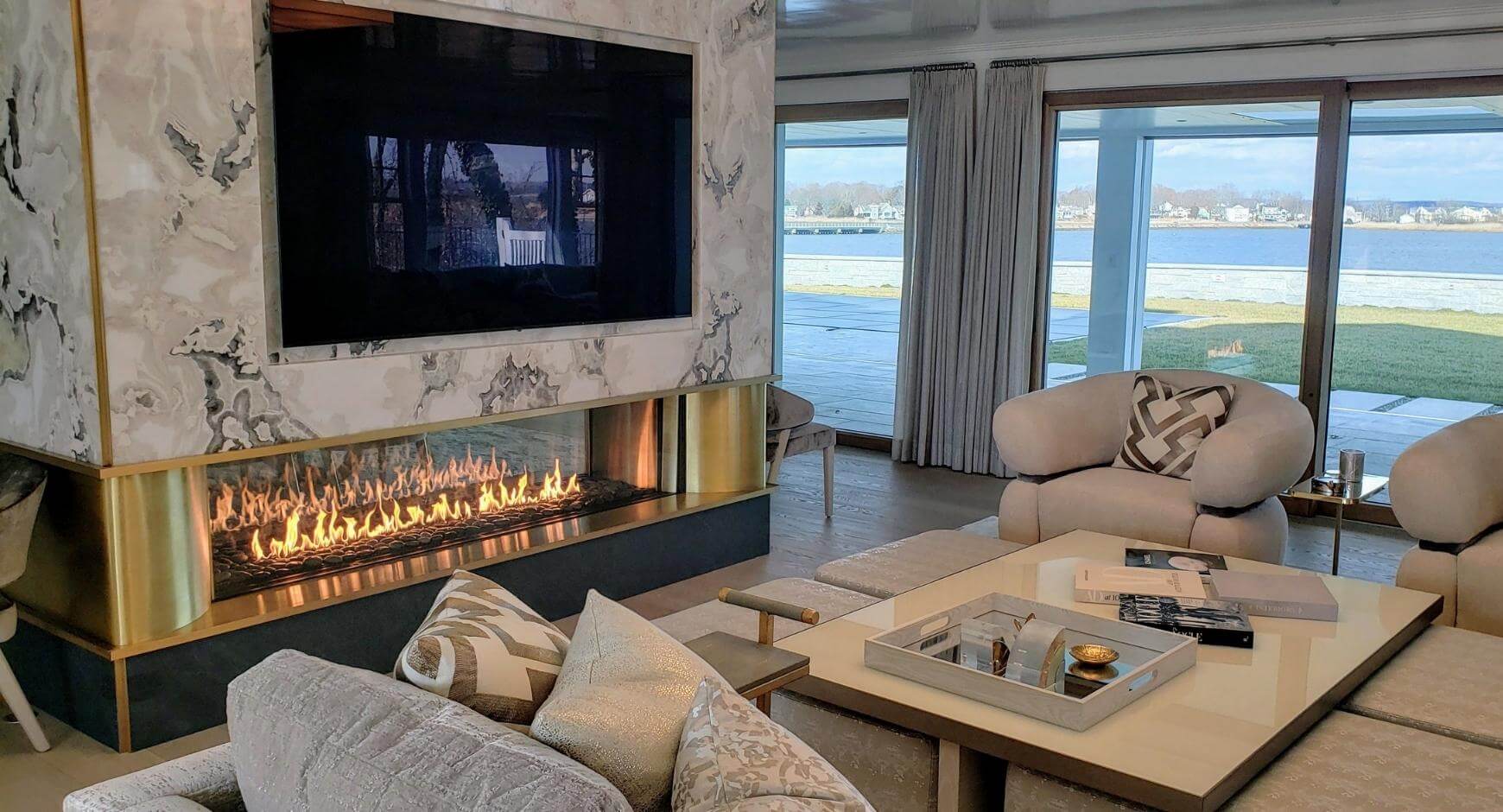 Luxurious living room with marble fireplace, modern gas flames, plush seating, and large windows overlooking water.