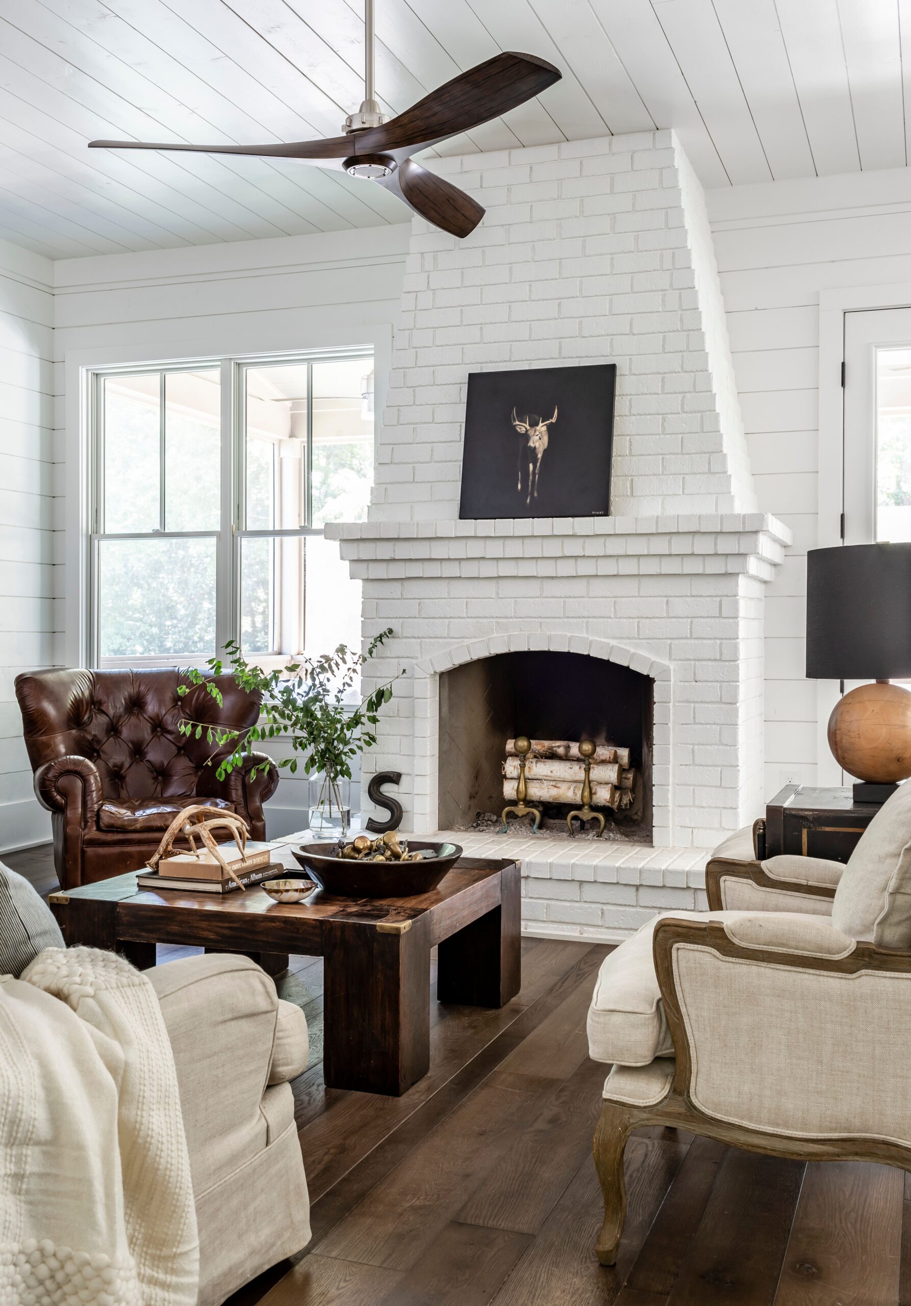 Cozy farmhouse living room with white brick fireplace, rustic wood coffee table, leather armchair, and neutral upholstered chairs.