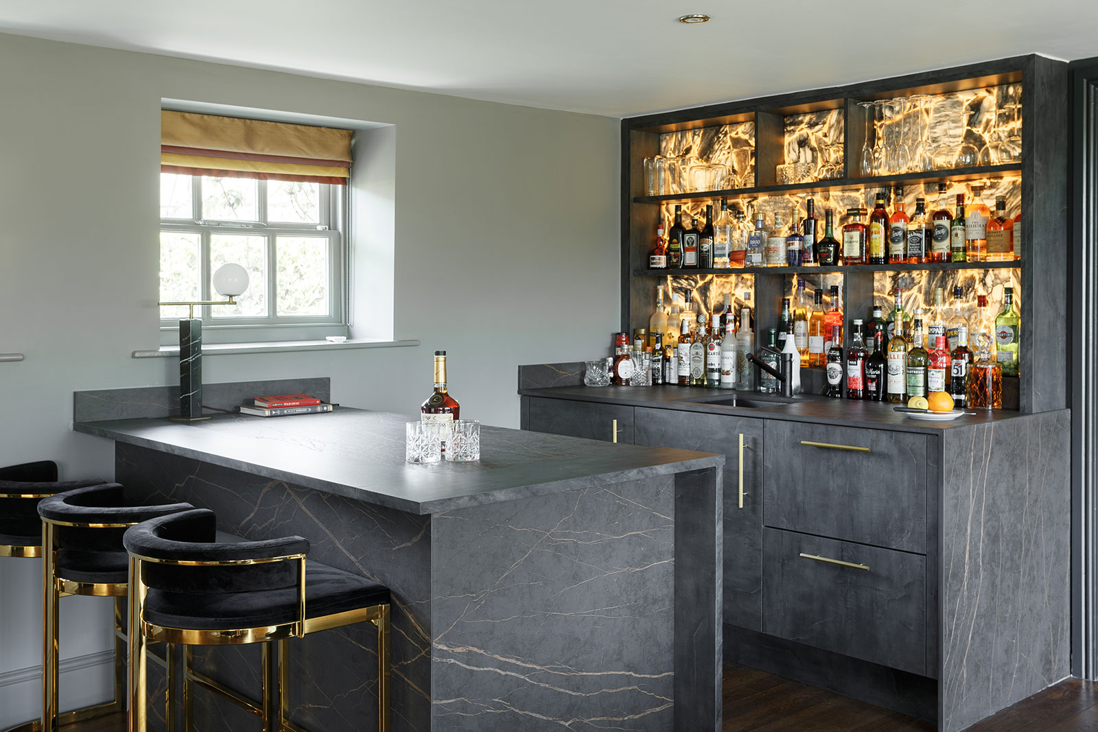 luxurious modern home bar with grey marble counters, gold and black bar stools, and illuminated liquor shelves