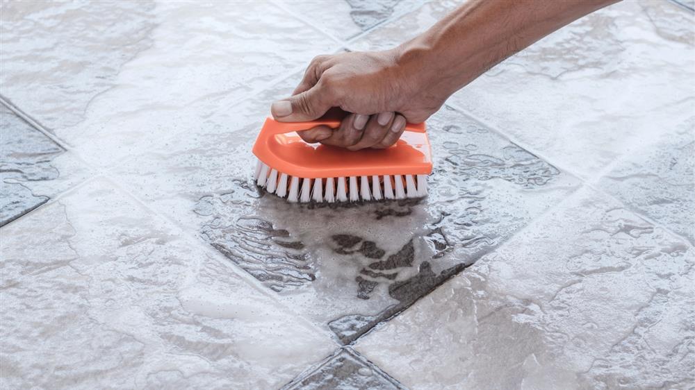 Hand scrubbing soapy water on ceramic floor tiles with an orange cleaning brush.