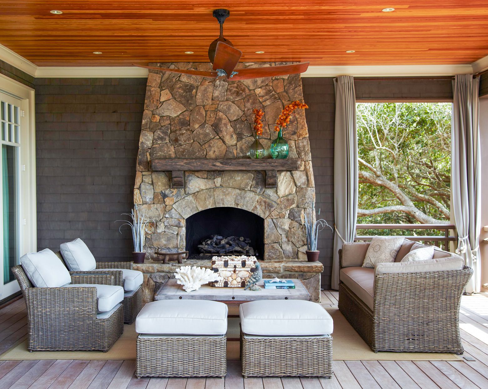 Covered outdoor patio with rustic stone fireplace, wicker furniture, wood ceiling, and serene tree views.
