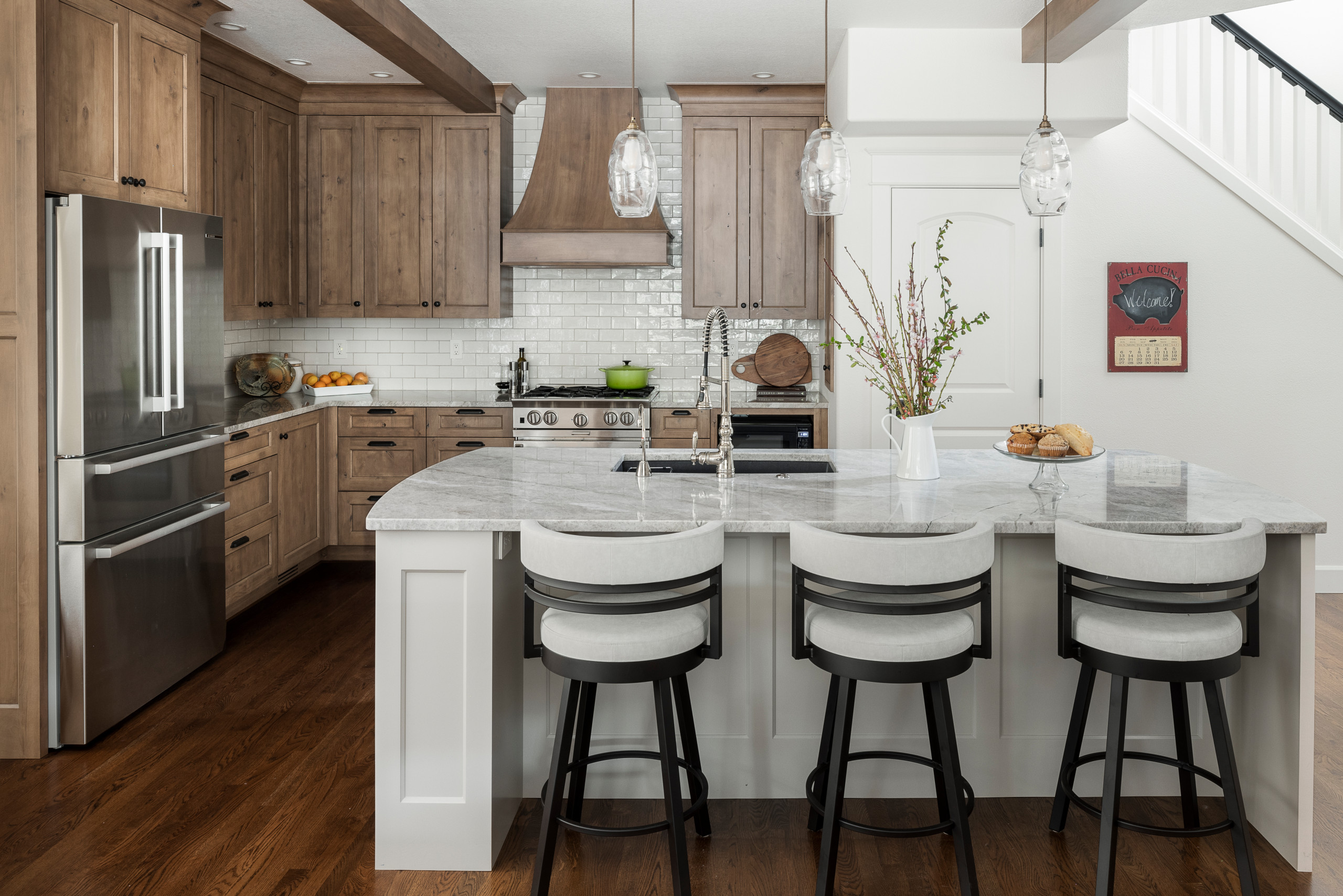 A cozy kitchen with rustic wood cabinetry, white subway tile backsplash, and a marble countertop island. Stainless steel appliances complement the warm tones, while pendant lights and upholstered stools add a modern touch.