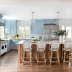 A spacious kitchen with soft blue cabinetry, a large wooden island, and white upholstered bar stools. Stainless steel appliances and pendant lights add a modern touch, while natural wood flooring enhances warmth.