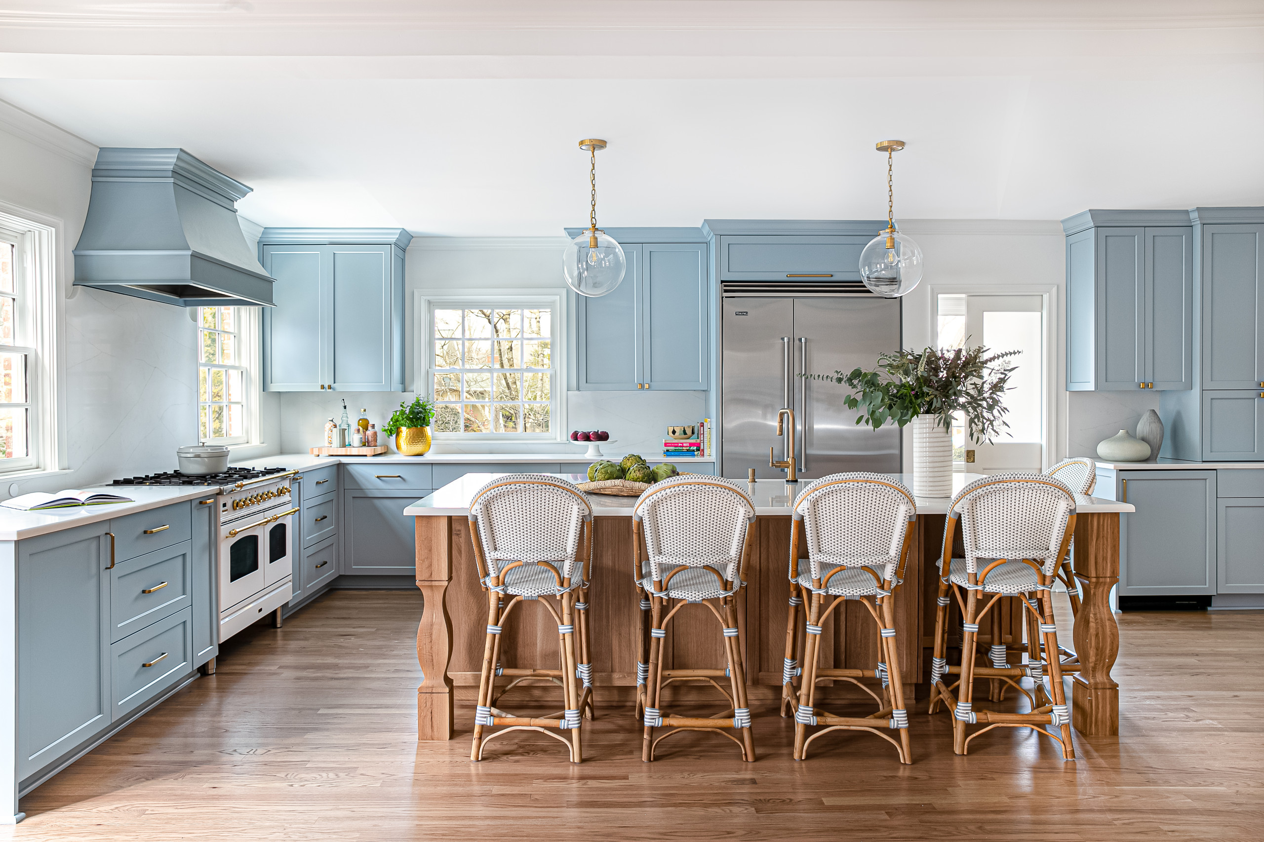 A spacious kitchen with soft blue cabinetry, a large wooden island, and white upholstered bar stools. Stainless steel appliances and pendant lights add a modern touch, while natural wood flooring enhances warmth.