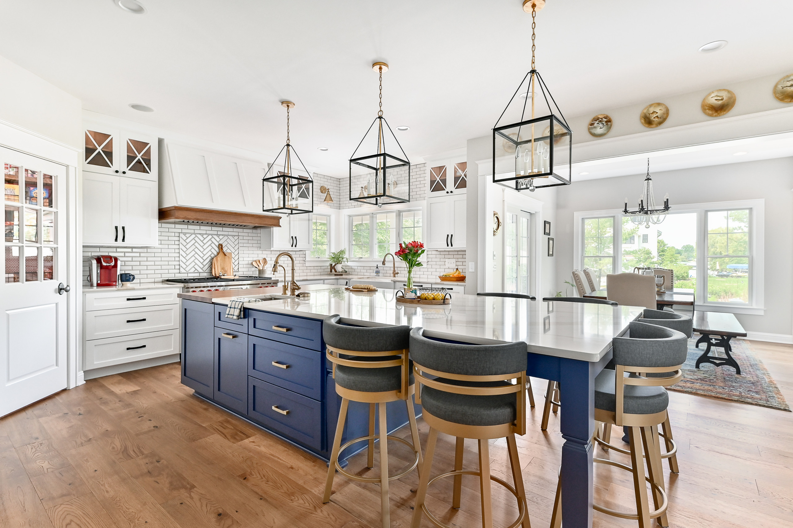 A spacious kitchen with a navy-blue island, white cabinetry, and hardwood flooring. Pendant lights in geometric frames hang above the island, surrounded by cushioned bar stools. The kitchen features a subway tile backsplash, stainless steel appliances, and a view of the dining area through large windows.