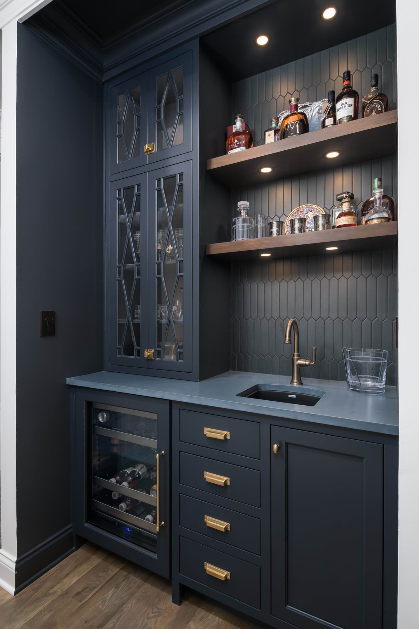 Modern home bar with navy blue cabinetry, glass shelves, and built-in mini fridge.