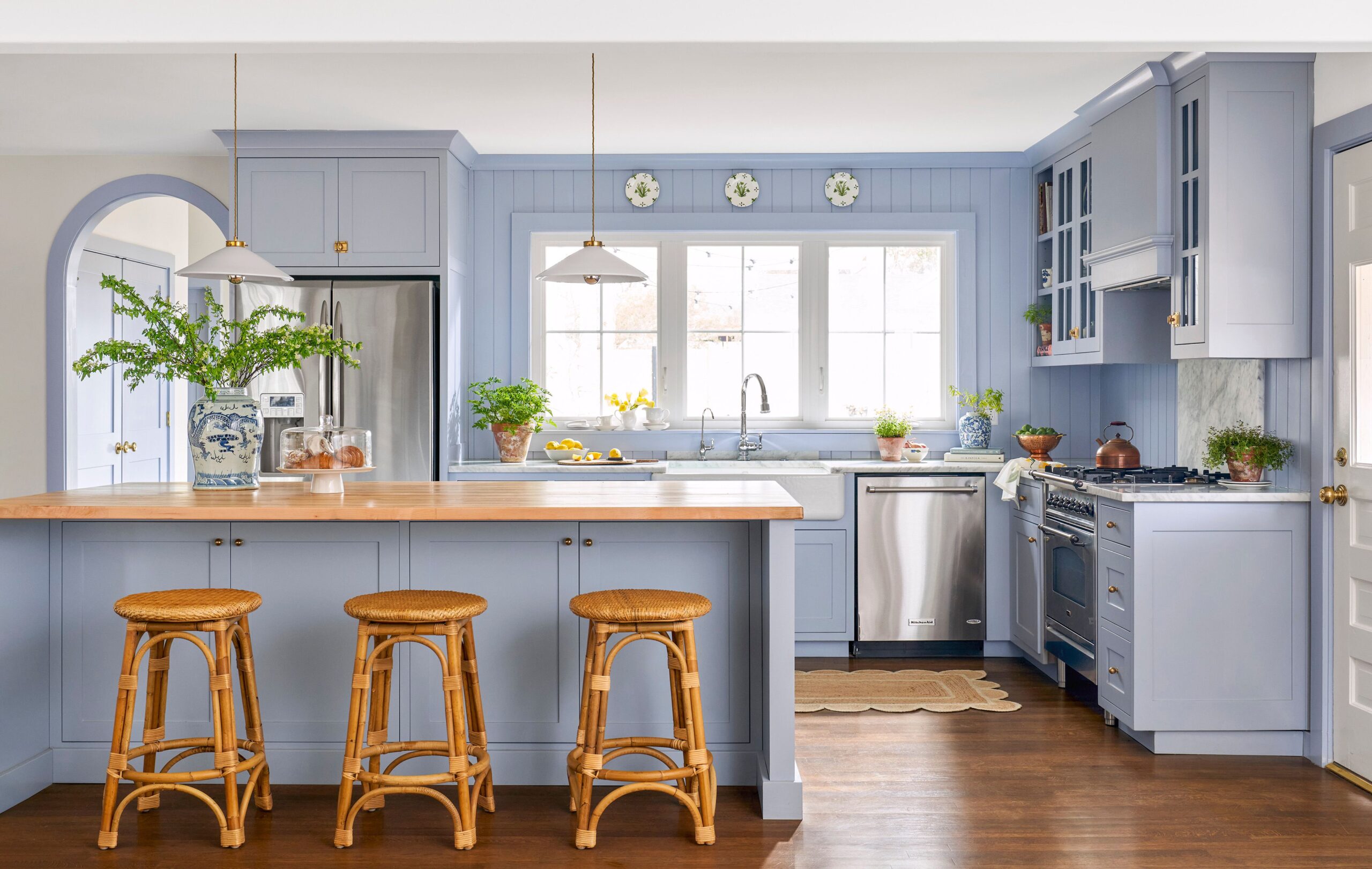 A bright kitchen featuring soft blue cabinetry, warm wood countertops, and rattan stools. Stainless steel appliances add a modern touch, while decorative accents and greenery bring charm and freshness to the space.