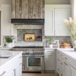 A modern farmhouse kitchen featuring white cabinetry, a rustic wood range hood, and stainless steel appliances. The backsplash includes herringbone tiles surrounded by subway tiles, while the countertops are white quartz. A wooden cutting board with fruit and decorative plants adds warmth to the space.