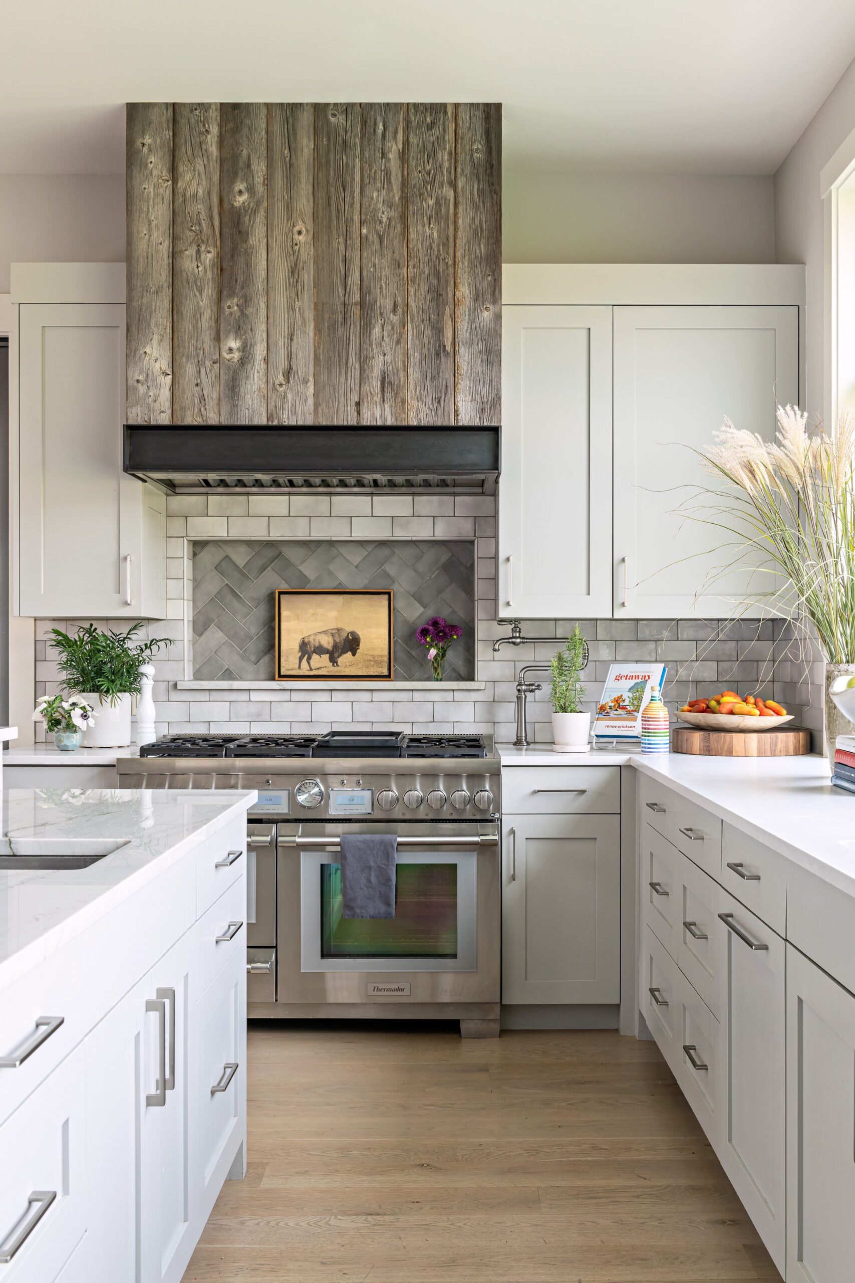 A modern farmhouse kitchen featuring white cabinetry, a rustic wood range hood, and stainless steel appliances. The backsplash includes herringbone tiles surrounded by subway tiles, while the countertops are white quartz. A wooden cutting board with fruit and decorative plants adds warmth to the space.