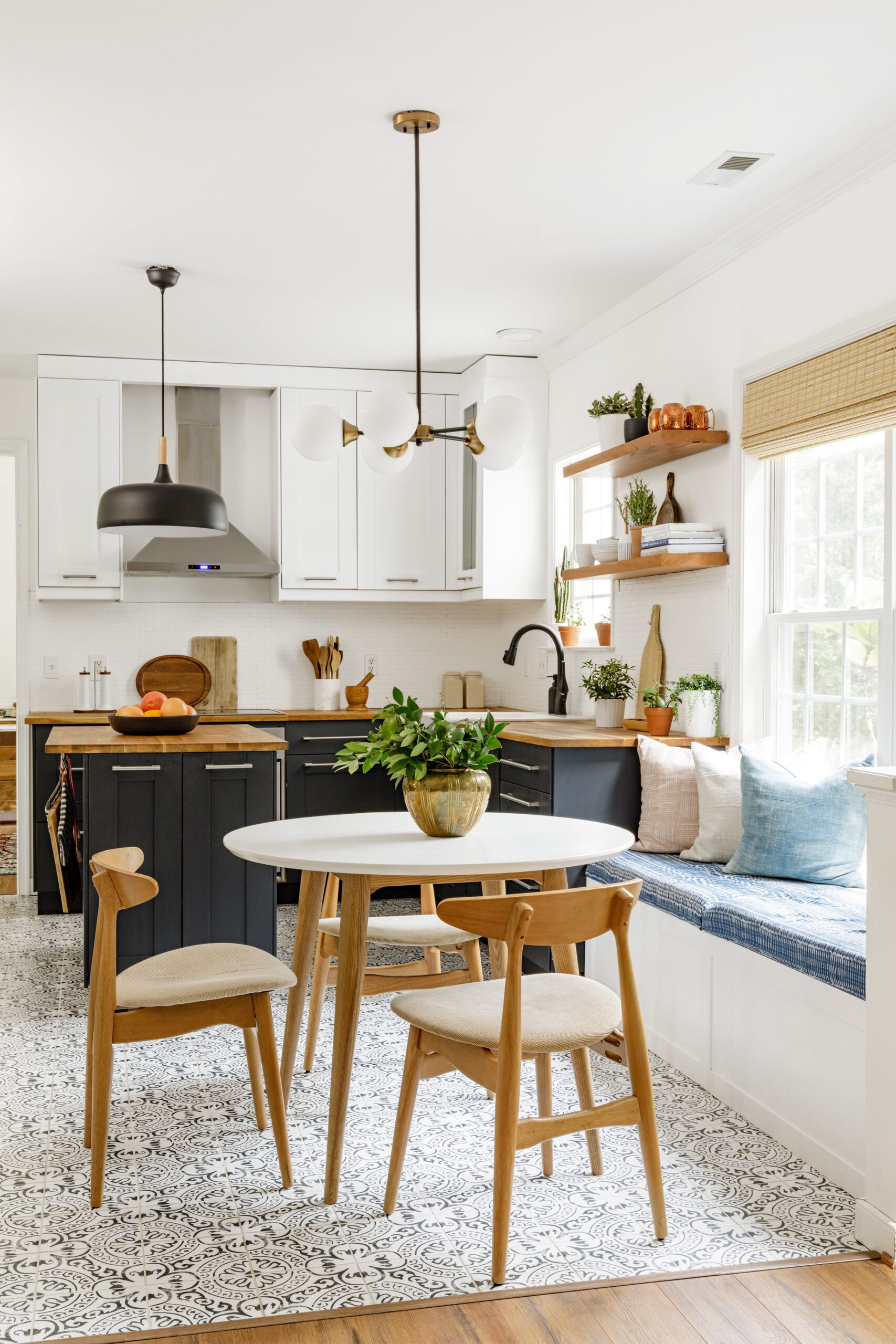 A cozy kitchen with navy-blue lower cabinets, white upper cabinets, and wooden accents. A round dining table with wooden chairs sits on patterned tile flooring, complemented by a built-in bench with cushions near a bright window.