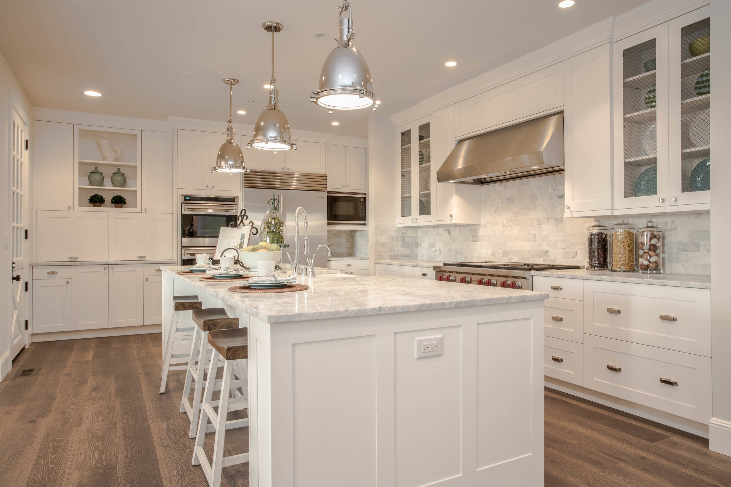 A modern farmhouse-style kitchen featuring white cabinetry, a marble island, stainless steel appliances, and wooden bar stools. Pendant lights hang above the island, while the hardwood flooring adds warmth to the space.