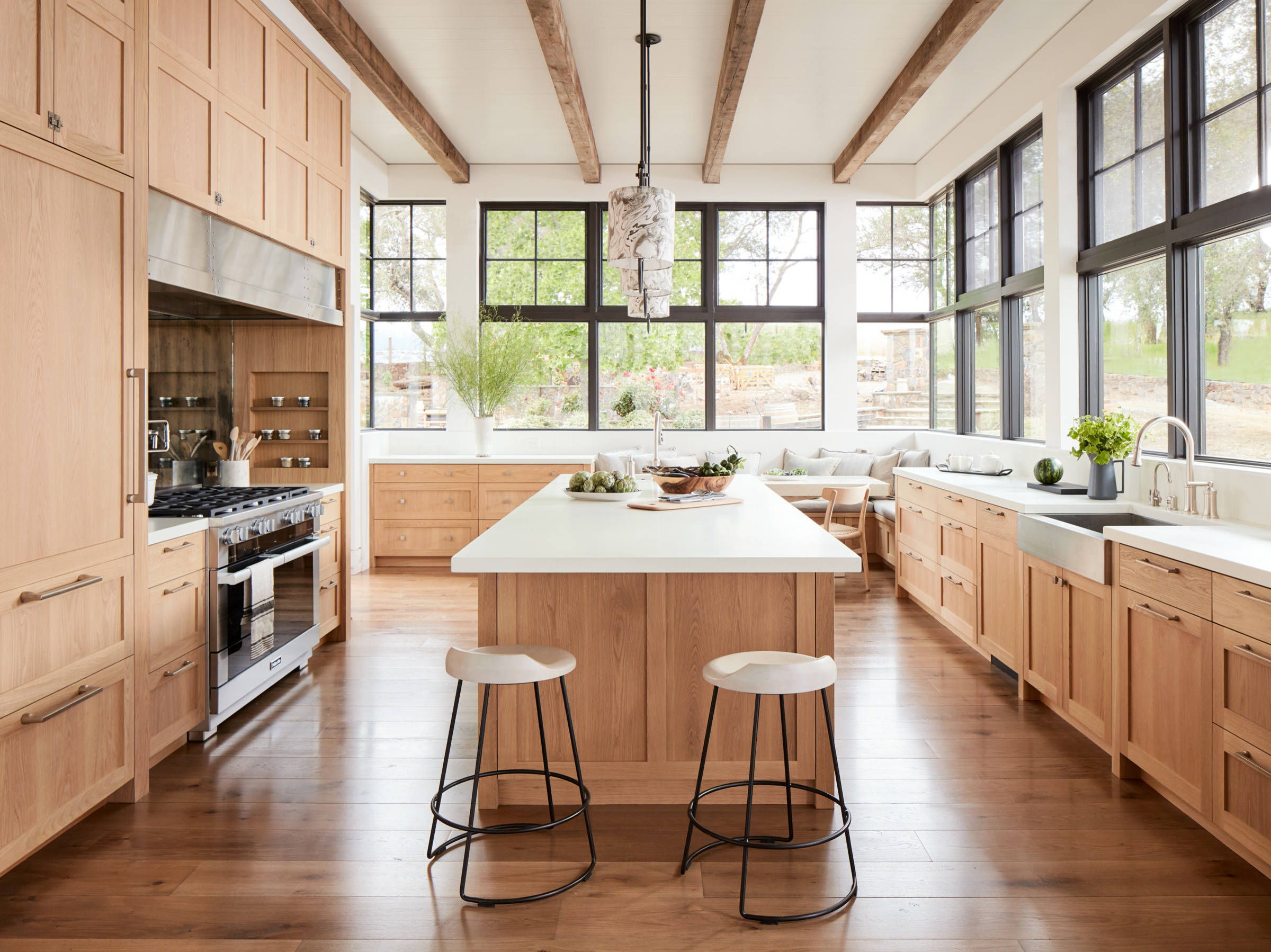 A bright and airy farmhouse-style kitchen with natural wood cabinetry, white countertops, exposed wooden beams, and large black-framed windows. The space includes a central island with stools and a cozy breakfast nook.