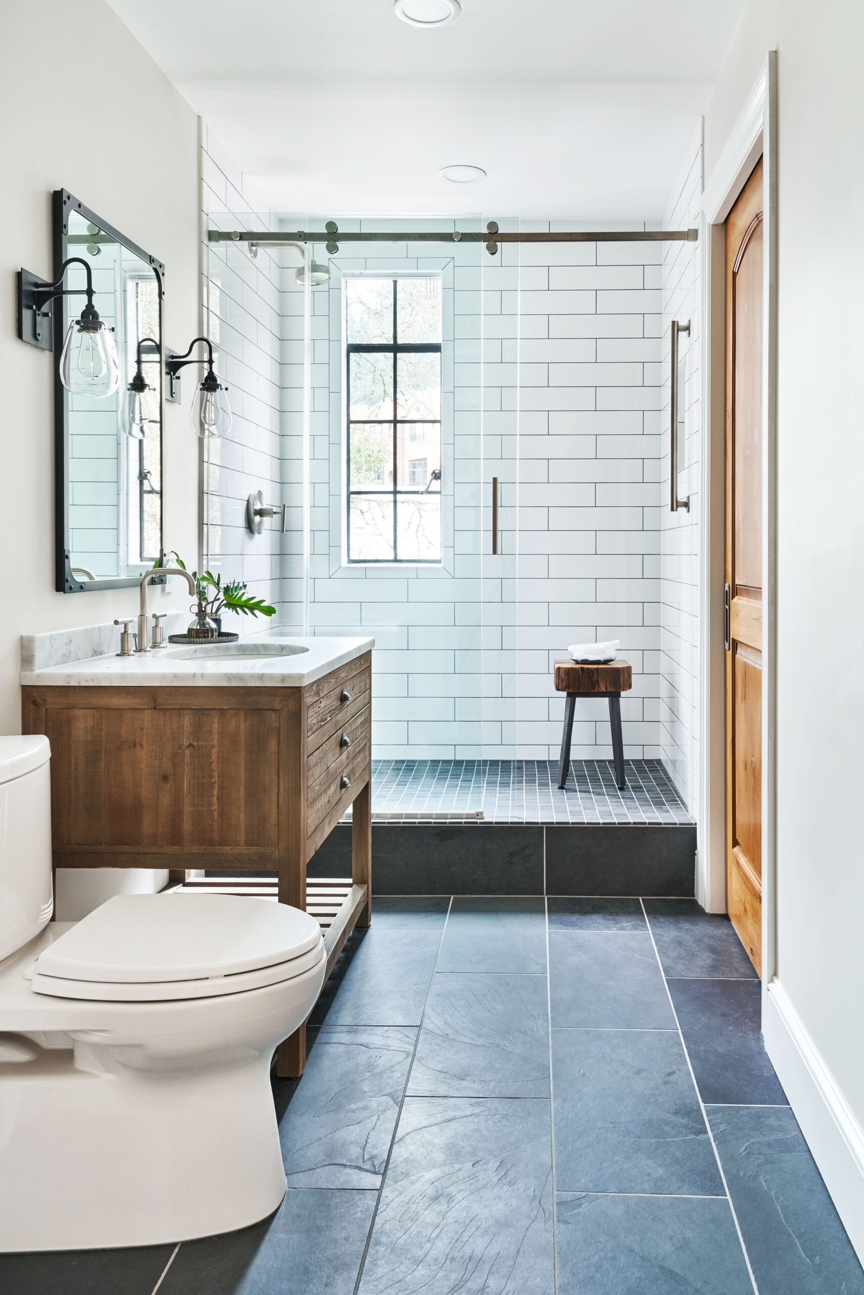 A modern bathroom featuring a white subway tile shower, a wooden vanity with a marble top, and sleek black tiles on the floor. There is a glass shower door, a small wooden stool, and a window allowing natural light.